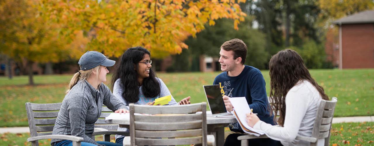 Students sitting at an outdoor table conversing with fall foliage in the background.