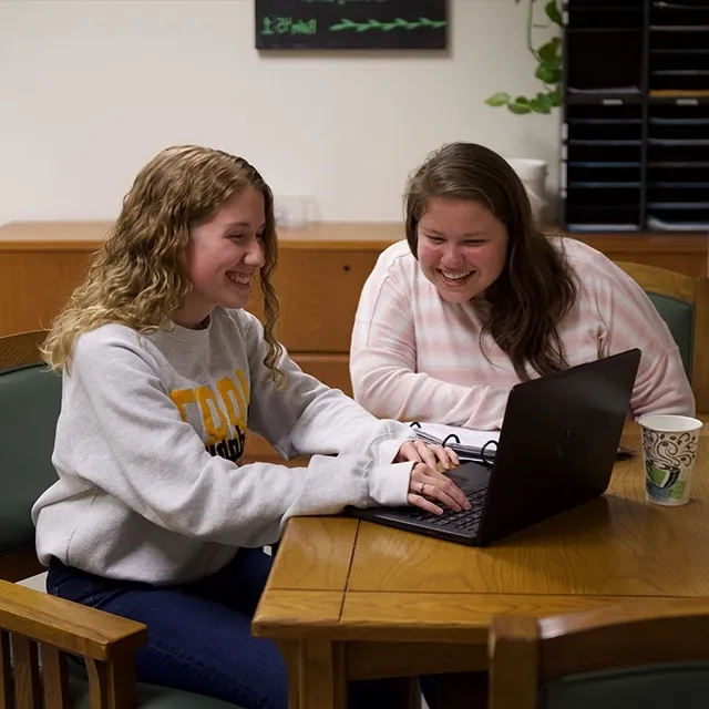 Female tutor helping a female student seated at a table working on a laptop computer.